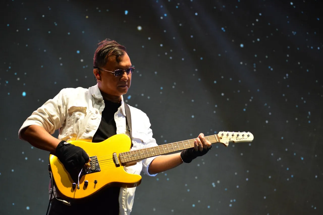 Ari passionately playing a bright yellow electric guitar on stage, wearing a white shirt and black gloves against a dark, starry backdrop with dramatic spotlighting.